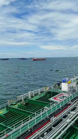 The video shows a wide shot of a body of water with several ships and barges visible. In the foreground, a large green and white vessel is docked, with its deck structure and pipes clearly visible. In the midground and background, several other large ships, some red and some dark, are anchored or moving slowly in the water. The sky is partly cloudy with a mix of blue and white. The water appears calm. The camera angle is high, looking down onto the scene from a higher vantage point, likely from another ship or a structure.