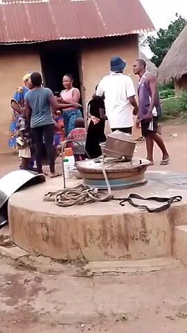 A group of people are gathered around a well in a dirt courtyard. The well is made of concrete and has a metal bucket and rope on top. There is a house with a corrugated metal roof in the background. The people appear to be talking and interacting with each other. The scene suggests a rural or village setting.