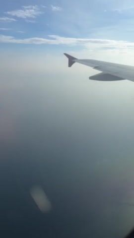 The video shows an aerial view from an airplane window. The right wing of the plane is visible in the foreground, with a red tip. Below the wing, a hazy expanse of water or land can be seen, shrouded in a light mist or cloud cover. The sky above is a clear blue with scattered white clouds. The scene is static, with no discernible movement other than what might be attributed to the plane's flight.