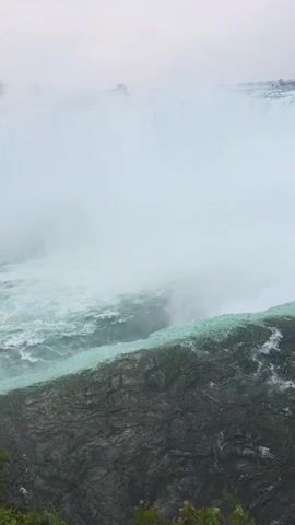 A POV shot looking down at the edge of a waterfall. The water is turquoise and frothy as it cascades over the edge. The rock face is dark and jagged. Mist from the waterfall obscures the view in the distance.