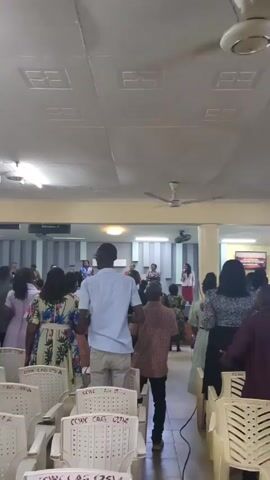The video shows a church service in progress. A large group of people, mostly adults, are standing in a large room with rows of white chairs. A group is assembled on a raised platform, which seems to be the stage area. They appear to be singing and leading the congregation. Some of the attendees are clapping. The room is well-lit with a low ceiling and several ceiling fans. A camera on a tripod is present, recording the event. The overall setting indicates a regular church service.