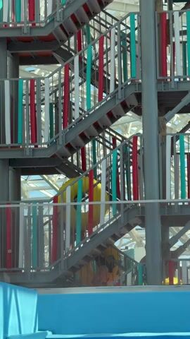 The video captures a multi-level metal staircase, likely part of an indoor water park in Kogalym, Russia. The staircase features striking railings composed of vertical bars in red, white, and turquoise, creating a colorful and dynamic visual. Several individuals, appearing to be children or young adults, are seen ascending the stairs. Each person carries a bright yellow inflatable tube, indicating they are preparing to use a water slide or similar attraction. One person is visible on a lower flight of stairs, while another is higher up the structure. In the background, a complex framework of metal and what appears to be a glass or translucent roof suggests an expansive indoor facility. A vibrant blue pool edge or structure is visible at the bottom of the frame. The scene depicts a recreational activity in a brightly lit, modern setting. There is no visible text.