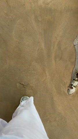The video shows a first-person perspective of someone walking on a sandy beach. The person is wearing white pants and sneakers. The sand is wet and appears to have small pebbles and shells on it. There are also footprints in the sand, indicating that others have walked on the beach. The camera pans slightly, revealing a large rock covered in green algae. The overall environment appears to be overcast.