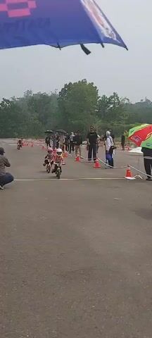 The video shows an outdoor children's bicycle race on an asphalt track. Several young children on small bicycles, wearing helmets and protective gear, are participating. Spectators, some holding umbrellas, are visible along the sidelines. An ambulance and medical personnel are present at the scene, suggesting a safety precaution. The weather appears overcast, possibly indicating rain. The race is demarcated by orange cones and a white line across the track, possibly a start or finish line. The overall atmosphere suggests a community sporting event for young children.
