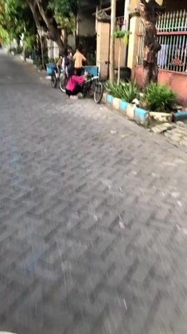 A short video shows two children on bicycles on a paved street. The first child, a girl wearing a pink shirt and black shorts, is kneeling next to a bicycle. The second child, a boy wearing a white and black striped shirt, is on a bicycle behind her. A person with dark hair and a white shirt is visible in the background, and another person with dark hair and a light-colored shirt is standing. The street is lined with trees and buildings. The camera then shows a close-up of a motorcycle's speedometer, indicating a speed of approximately 20 km/h. The video appears to be filmed from the perspective of someone riding a motorcycle.