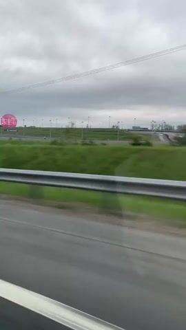 The video shows a view from a moving car, driving on a road with a metal guardrail. On the other side of the guardrail is a grassy field, dotted with evenly spaced lampposts. In the distance, atop a small hill, there is a large, illuminated pink sphere. The sky is overcast and grey. The car's side mirror is visible in the lower left corner of the frame. The general scene is unremarkable, capturing a view of the landscape from a vehicle on the road.