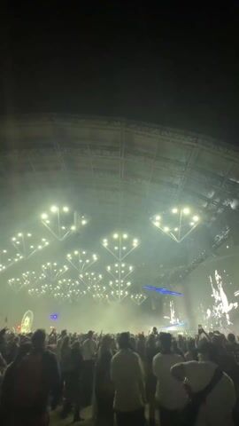 The video shows a large crowd of people at a concert, at night, indoors.  The ceiling of the venue is high and has a complex geometric pattern, illuminated by bright white spotlights.  Blue laser beams are also visible. In the distance, beyond the crowd, is a Ferris wheel. Most people in the crowd are looking towards the stage, with some raising their hands or holding up their phones.  The overall atmosphere is dark and energetic.