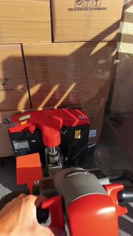 A first-person perspective shot shows a person's hands operating a red electric pallet jack. The pallet jack is positioned in front of a stack of cardboard boxes wrapped in plastic. The boxes have "DIKKAT KIRILIR!" (Caution Fragile!) printed on them. The pallet jack is moving forward, about to lift the boxes. The environment appears to be outdoors, with sunlight casting shadows on the boxes. The ground is paved with brick-like material.