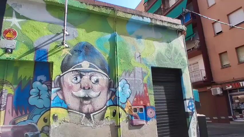 This is a slow pan of a building exterior covered in vibrant graffiti. On the left, a multi-paneled portrait of a face is painted, with various sections of different colors and textures. In the center, a large cartoonish face with a mustache and a hat is depicted, with cartoonish clouds and industrial imagery around it. To the right, a dark metal door is visible, also with some graffiti. In the background, parts of apartment buildings with balconies and awnings can be seen. The scene appears to be an urban environment on a sunny day.