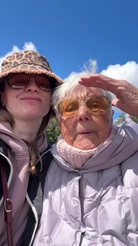 A woman in a leopard print bucket hat, sunglasses, and a pink sweatshirt stands next to an older woman with white hair, amber tinted sunglasses, and a light purple jacket. Both are outdoors with a blue sky and scattered clouds visible in the background.  The younger woman is taking a selfie and smiles, then kisses the older woman on the cheek. The older woman looks directly at the camera with a neutral expression. The setting appears to be a park or outdoor event. There are trees and street lights in the background. Music plays in the background.