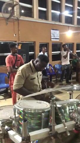 The video shows a live musical performance inside a room, possibly a church or a community hall, with warm-toned walls and multiple windows. The focus is on a drum set being played by a man in a khaki shirt. Behind him, another man in a red shirt stands near a keyboard player and sings into a microphone. The keyboard is a Yamaha model. There are other people in the background, some standing and clapping. The room is illuminated by overhead lights and there's a ceiling fan rotating. Chairs are scattered around the room, some blue plastic ones visible near the drummist. The video's perspective is from the front of the drum set, providing a clear view of the drummer and surrounding musicians.