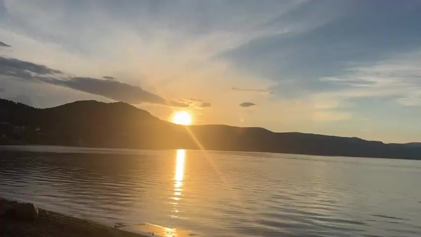 The video captures a sunset over a large body of water, likely a lake, viewed from a shoreline. The sun, a bright orb, is partially obscured by rolling hills in the background, casting a golden glow and reflecting a shimmering path across the water's surface. The sky is a gradient of blues and grays, with scattered clouds, some of which appear to be tinged with the sunset's warm hues. On the left side of the frame, the shoreline is composed of gravel and sparse grass, with a few large rocks and a small concrete structure or pier. Further inland, nestled against the hills, a small village or settlement is visible, with houses appearing as dark silhouettes against the brighter sky. The water exhibits gentle ripples, suggesting a calm atmosphere. The overall impression is one of natural beauty and tranquility during the transition from day to evening. No people or animals are clearly discernible in the footage.