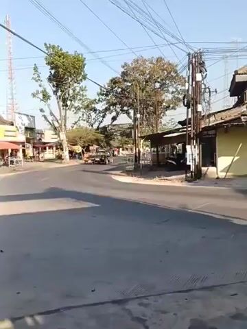 A man on a motorcycle is turning at an intersection in a rural or semi-rural area. There are several trees, power lines, and buildings visible. The sky is clear and blue. The scene appears to be during daylight hours. The road is paved and there are some shops and stalls on the side of the road.