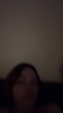 A blurry video shows two people in a dimly lit room. The person in the foreground is a female with dark hair, wearing a strapless top and a necklace. She looks at the camera and sticks out her tongue. To her right is a male with dark hair, partially visible. The background is dark and indistinct.