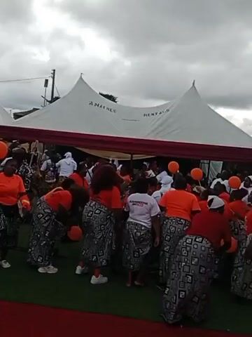 A wedding or large event is taking place outdoors under large white tents. The ground is covered with a red carpet and green turf. Several people are dressed in bright orange tops and patterned skirts. Some individuals are holding orange balloons. There are a few individuals in white shirts. A woman in a blue shirt with a logo is carrying a green tray. The atmosphere appears festive.