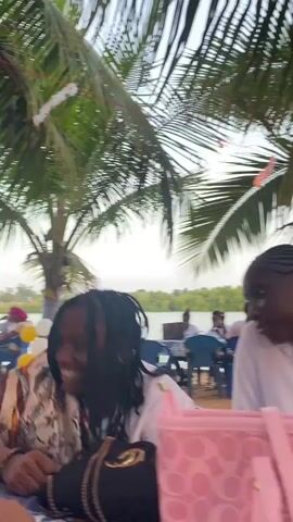 The video depicts an outdoor event, possibly a party or gathering, held on a beach under palm trees. The scene includes several people, some seated at tables with floral tablecloths, and others standing or walking around. A DJ booth is set up near a palm tree, and a man is operating the equipment. The environment is sunny and relaxed, with water visible in the background. A pink patterned bag is in the foreground, partially obscuring the view. The people are of various ages and attire, and the general atmosphere seems festive and social.