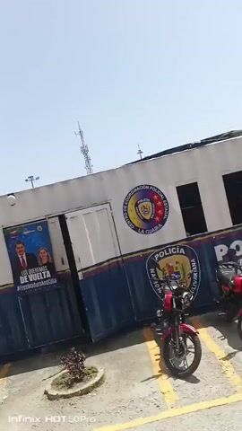 This video captures street scenes in Maiquetía, Venezuela, on a bright, sunny day. The footage begins by focusing on a building with a banner that reads "Ministerio del Poder Popular para Relaciones Interiores Justicia y Paz," translating to "Ministry of Popular Power for Interior Relations, Justice, and Peace." The building also displays a circular emblem with "Gobernación Bolivariana de La Guaira" within it. A poster featuring political figures is visible, along with two motorcycles parked in front of the building.

The camera then pans to show a white pickup truck with "CONDUSEGURO" printed on its side, parked near a dense green bush. Further along, a red motorcycle is parked in front of a mural depicting various figures. The scene shifts to a tree-lined street where two individuals are standing. One person, wearing a red dress, is looking towards the camera, while another, in dark clothing, is partially obscured by the tree.

The video continues with shots of the street, showing parked cars, including a red sedan, and more traffic cones. In the background, various buildings, fences, and communication towers are visible. The bright sunlight suggests it is daytime, and the clear sky indicates fair weather. The overall atmosphere appears to be of everyday street activity in a Venezuelan city. The visible text primarily relates to governmental entities and a public safety program.