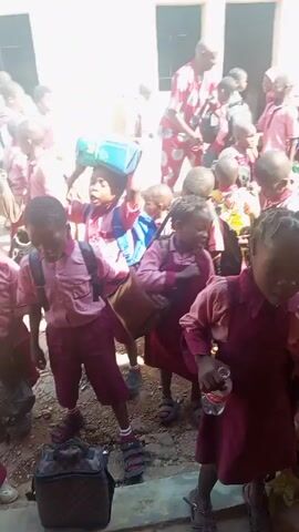 The video shows a group of children, likely students, in school uniforms. The uniforms vary in color and style, with some wearing maroon outfits and others in pink. The children are standing and moving around in what appears to be a school hallway or outdoor covered area. Some have backpacks and lunchboxes. In the background, there's a yellow van with visible text. An adult, possibly a teacher, is seen among the children. There is some shouting happening too.