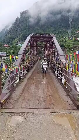A person on a scooter rides across a bridge adorned with prayer flags. The bridge is a metal truss structure, painted red, and it spans over a river. The road on the bridge appears wet and somewhat muddy. On either side of the bridge, there are metal railings also decorated with prayer flags. Beyond the bridge, there are steep, lush green hills with some buildings visible on the slopes, partially obscured by mist and clouds. A tall communication tower stands on one of the hills. The weather appears overcast and possibly rainy. Another person is visible walking further down the bridge.