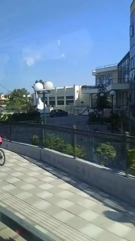 The video captures a street scene with a clear blue sky. In the foreground, a pedestrian walkway with a checkered pattern is visible. A person in a pink shirt is riding a bicycle on this walkway, moving from left to right. Behind the walkway, there is a fence and some greenery. Further in the background, a modern building with large windows and some external lighting fixtures is present. Several cars are on the road, including a silver sedan, a white pickup truck, and a dark-colored car. There appears to be a roundabout with some landscaping in the middle, including rocks and a small water feature or fountain. Trees and residential buildings are also visible in the distance. The lighting suggests it is daytime, likely in the morning or afternoon, given the shadows.