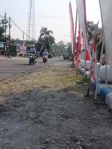 The video shows a roadside scene with several motorcycles and a car traveling on a paved road. The road appears to be in a rural or semi-rural area, with trees and buildings visible in the background. There are also several Indonesian flags displayed along the side of the road, suggesting a national holiday or celebration. The ground on the side of the road is unpaved and appears to be made of gravel and dirt. The sky is clear and sunny.