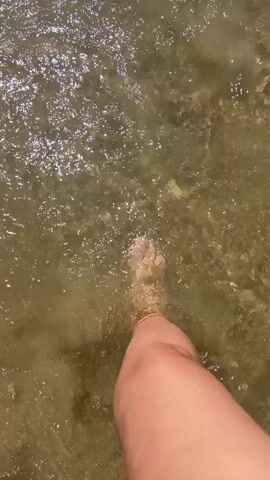 A first-person perspective video shows feet walking through shallow, murky water. The water is disturbed by the movement, creating ripples and splashes. The sunlight reflects off the water's surface, causing it to shimmer. The bottom of the body of water appears to be covered in sand or small pebbles. The person is wearing no shoes, and their feet are visible as they move through the water.