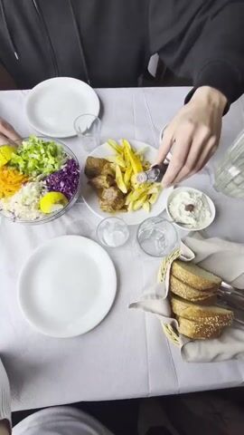 The video captures a dining table covered with a white tablecloth, where two people are preparing to enjoy a meal. A person wearing a black hoodie is using a tool that appears to be a small bottle opener or serving utensil to add something to a glass of water. The table is set with two empty white plates, a bowl of colorful salad with ingredients such as shredded lettuce, carrots, and red cabbage, a plate of meat and fries, a small dish of white dip, and a basket of sliced bread. There are also small glass cups with ice and a glass carafe visible on the table.