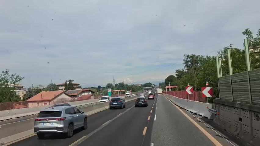 The video shows a view from a vehicle driving on a highway during the day. Several other vehicles are on the road. The highway has multiple lanes and is bordered by concrete barriers and greenery. Buildings and a line of trees are visible on the left side of the road. In the distance, snow-capped mountains can be seen on the horizon, and a high-pitched beep is heard. The sky is cloudy.