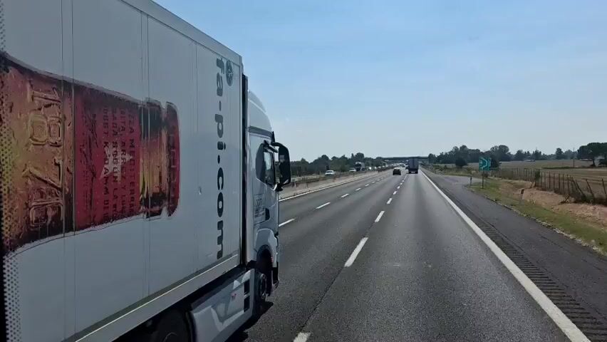 A first-person perspective video shows a truck driving on a highway. The truck has a large image of a beer bottle on its side, with the words "Estrella Barcelona" visible. The highway is multi-lane with other vehicles in the distance. The weather appears to be sunny with a clear blue sky. The landscape alongside the highway consists of fields and trees.