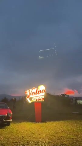 The video captures an outdoor scene at dusk. In the foreground, a red classic car is partially visible on the left. A tall, illuminated sign for "Volvo" with "Strecken & Sudsee 2019" beneath it stands prominently in the center of the frame, casting a yellow glow on the grass below. The sky is overcast with dark clouds, but there are pockets of vibrant orange and red light, suggesting a sunset. In the middle ground, several people are walking across a grassy field. One person appears to be carrying something over their shoulder, and another is pushing a bicycle. In the distance, hills and trees are silhouetted against the colorful sky. High up in the cloudy sky, a formation of lights is visible, resembling a bus or a train. The overall atmosphere is serene and somewhat moody due to the twilight conditions and the striking sky colors.