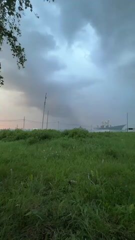 The video shows a wide shot of a field of green grass under a cloudy sky. In the distance, there are some trees and a building complex. A rain shaft is visible in the middle ground, extending from the clouds to the ground. The overall mood is somber and atmospheric, suggesting an approaching storm. The camera appears to be held steady, with no discernible movement or panning. The lighting is dim and diffused, consistent with overcast weather.