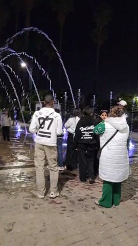 At night, a group of people are gathered around a lighted fountain. The fountain has multiple jets that shoot water in arcs and straight streams. Some of the jets are illuminated with purple light. Several people are walking near the fountain. The camera is moving around as it captures the scene. The ground around the fountain is wet. Palm trees are visible in the background.