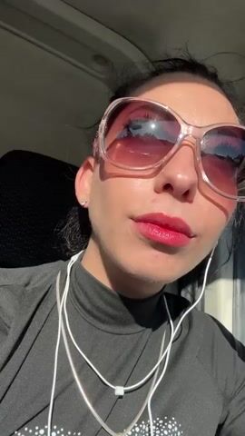 A woman with dark hair pulled back into a bun, wearing large, round, light-colored sunglasses, a black turtleneck shirt, and a silver chain necklace, is inside what appears to be a vehicle. She is looking directly at the camera and singing along to music playing through white wired earphones. Her lips are painted a bright red. The sunlight streams into the vehicle from her side.