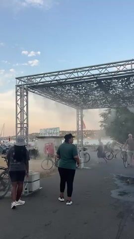 The video shows an outdoor scene with a large metal structure overhead, possibly a stage or canopy, with a decorative pattern on its surface. Misting fans are attached to the structure, creating a cool, misty environment. Several people are present, some walking or standing, while others are with bicycles. In the foreground, two individuals are visible from behind, one wearing a striped hat. Further back, a person in a green shirt stands facing away from the camera. A man in shorts and a t-shirt is seen holding a bicycle, looking towards the camera. The background suggests a waterfront or park area with trees, buildings, and a body of water, possibly a lake or river. The lighting indicates it is either early morning or late afternoon, with soft, warm light. The overall atmosphere appears to be one of leisure or an outdoor event, with the mist providing a welcome respite from the heat.