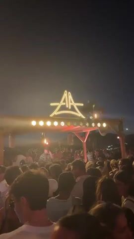 The video shows a crowded outdoor event at night, likely a festival or concert. A large group of people are gathered in front of a raised stage or DJ booth. Above the stage, a neon sign with the letters "AA" in a stylized font is illuminated. The sign is supported by a wooden structure adorned with string lights. A DJ or performer is visible on the stage, surrounded by equipment. The atmosphere appears lively and energetic, with the crowd densely packed and looking towards the stage. The sky is dark, indicating it is nighttime. The lighting is dim, with spotlights and the neon sign providing the primary illumination. Smoke or haze is present in the air, contributing to the stage lighting effects.
