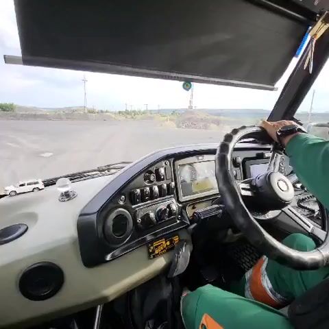 This video shows a first-person perspective of someone driving a vehicle, likely a large truck or bus, on an unpaved road. The driver's hands are visible on the steering wheel, and they are wearing a green long-sleeved shirt and green pants with orange accents. The interior of the vehicle is visible, including the dashboard with various buttons and a screen displaying what appears to be navigation information. Outside the vehicle, the landscape is dusty and barren, with hills in the background and what appear to be power lines or industrial structures. The sky is overcast.