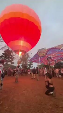 The video shows a crowd of people gathered in a muddy field with a colorful hot air balloon rising overhead. The balloon is primarily red, pink, purple, and orange, with the burner actively flaming to provide lift. To the right of the balloon is a large, colorful, patterned awning providing shade over part of the crowd. The people are milling about, watching the balloon. Several people are holding up their phones or cameras, seemingly taking photos or videos. One woman is kneeling in the foreground, taking a picture with a camera. The overall atmosphere suggests a festival or event. The sky is overcast and grey.