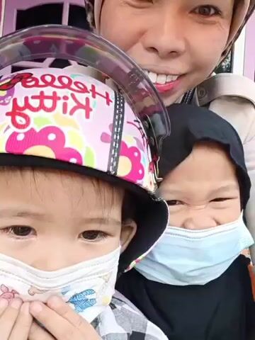 A close-up selfie-style video shows a woman and two young children. The woman is in the background, smiling broadly with her eyes slightly squinted. She is wearing a light-colored head covering. In the foreground, a child is on the left, wearing a helmet with a Hello Kitty design, a mask, and a plaid shirt. Another child is on the right, wearing a black head covering and a light blue mask. Both children are looking towards the camera. The environment appears to be outdoors, with a pink structure visible in the background. The video captures a brief moment of interaction between the family members.