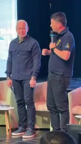 The video shows two men standing on a stage in front of a screen that reads “IMT”. The man on the left is white-haired and wearing a dark blue button-up shirt, dark pants, and gray sneakers. He has a bracelet on his right wrist and is holding a tablet in his left hand. The man on the right has short dark hair and is wearing a dark blue polo shirt with a yellow and blue logo on the left chest, dark pants, and a watch on his left wrist. He is holding a microphone in his right hand and gesturing with his left hand as he speaks. There is a light wood end table to the left of the men with a clear glass and a bottle on it. There is a pink upholstered chair to the right. The background is a dark curtain and the stage is lit.