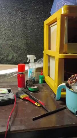 A static, medium-shot video focuses on a workbench littered with tools and materials. On the left side, a black digital multimeter with red and black wires is visible. To its right, several paintbrushes are laid out, along with a can of spray lubricant and a small container of what appears to be solder. A green bottle of glue and a white spray bottle are also present. In the background, a yellow plastic shelving unit holds various items, including what look like framed pictures or screens. A blue cup or container sits on the right edge of the frame. The workbench surface is dark and appears worn. In the far background, a dark, textured screen or netting is visible. A tripod with a weight attached to it stands in the middle background, with a speaker and a turquoise water bottle near its base.