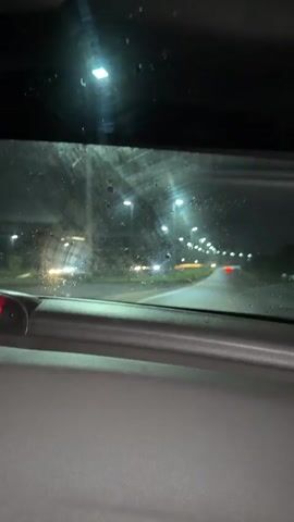 A nighttime drive through the rain, as viewed from inside a car. The wipers work periodically, clearing raindrops from the windshield. The road ahead is illuminated by streetlights, with the lights of other cars visible in the distance. The dashboard of the car is visible in the lower portion of the frame.
