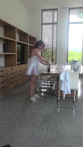 A woman with red hair, wearing a white short dress and white slippers, stands next to a wooden dining table in a bright room. She is pouring liquid from a dark bottle into a wine glass. Several other wine glasses are arranged on the table, along with a white gift bag and a white robe draped over a chair. The woman has tattoos visible on her legs. In the background, there are extensive wooden shelving units with a television, and large windows with blinds offering a view of greenery outside. The woman looks towards the camera and smiles.