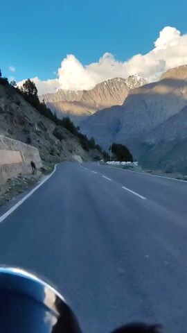 The video shows a first-person perspective of a person riding on a paved road through a mountainous region. The road curves to the left and ahead. The left side of the road features a steep, rocky hillside with scattered trees and vegetation. A low stone wall is visible along the edge of the hillside. The right side of the road opens up to a valley with more mountains in the distance. These distant mountains are snow-capped and illuminated by sunlight, contrasting with the shaded slopes closer to the road. The sky is blue with scattered white clouds. The bottom of the frame shows a blurry reflection, possibly from a vehicle's windshield or helmet. The overall scene depicts a scenic drive in a mountainous landscape, likely during daytime given the lighting conditions.