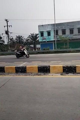 The video shows a road scene with a concrete curb painted with alternating yellow and black stripes. On the left side of the road, there are several buildings, palm trees, and other vegetation. There are also power lines and poles. Several motorcycles are parked along the side of the road. Two people are riding on one of the motorcycles. The sky is overcast.