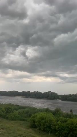 The video shows a wide river flowing through a landscape dominated by lush green trees lining its banks. The sky above is filled with heavy, dark gray clouds, suggesting an impending storm or overcast weather. The overall impression is one of a natural, somewhat moody environment. There are no visible people or man-made objects in the frame besides a small boat visible on the river in the distance.