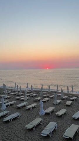 The video shows a beach at sunrise. The sun is a bright orange disk peeking over the horizon, casting a pink and orange glow across the sky and the water. The beach is covered in sand, and there are many rows of empty white lounge chairs and closed white umbrellas. The waves are gently lapping at the shore. The scene is peaceful and serene.