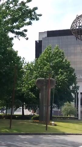 The video shows a street scene in front of a modern building with a glass facade. A large, spherical metal sculpture hangs from a wire in front of the building. The building has the words "ESTAÇÃO DE BRAGA" visible on its facade, indicating it is the Braga Station. There are trees and landscaping in front of the building, and cars are visible on the street. The sky is clear and blue. The video pans slowly from left to right, offering a wider view of the area.