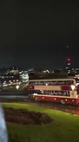At night, a red and white double-decker bus moves along a road parallel to a landscaped hill in a city.  The bus is well-lit with numerous passengers visible in the upper deck windows. In the background, city buildings are visible under a cloudy sky. Two red lights on a tall structure also punctuate the skyline. The camera briefly passes by a black metal fence.