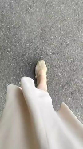 The video shows a first-person perspective of someone walking. The camera is pointed downwards at the feet and the ground. The ground appears to be asphalt or a similar paved surface, with some small debris, possibly leaves or pebbles, scattered on it. The person is wearing light-colored shoes and a long, flowing garment, possibly a dress or skirt, which partially obscures their legs and feet as they walk. The movement suggests walking at a steady pace. No other people or distinct objects are visible. The lighting appears to be natural, suggesting an outdoor setting.