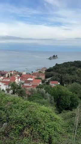 The video pans from an overview of a townscape with red-tiled roofs to a close-up of a small, rocky island. The town sits on a coast, surrounded by trees and vegetation. In the distance, the sea stretches to the horizon under a cloudy sky. Sailboats are visible in the water near the town. As the video zooms, the focus shifts to the rocky island, which features a small, simple structure with a red roof on its peak. The sea surrounding the island is calm and slightly rippled.